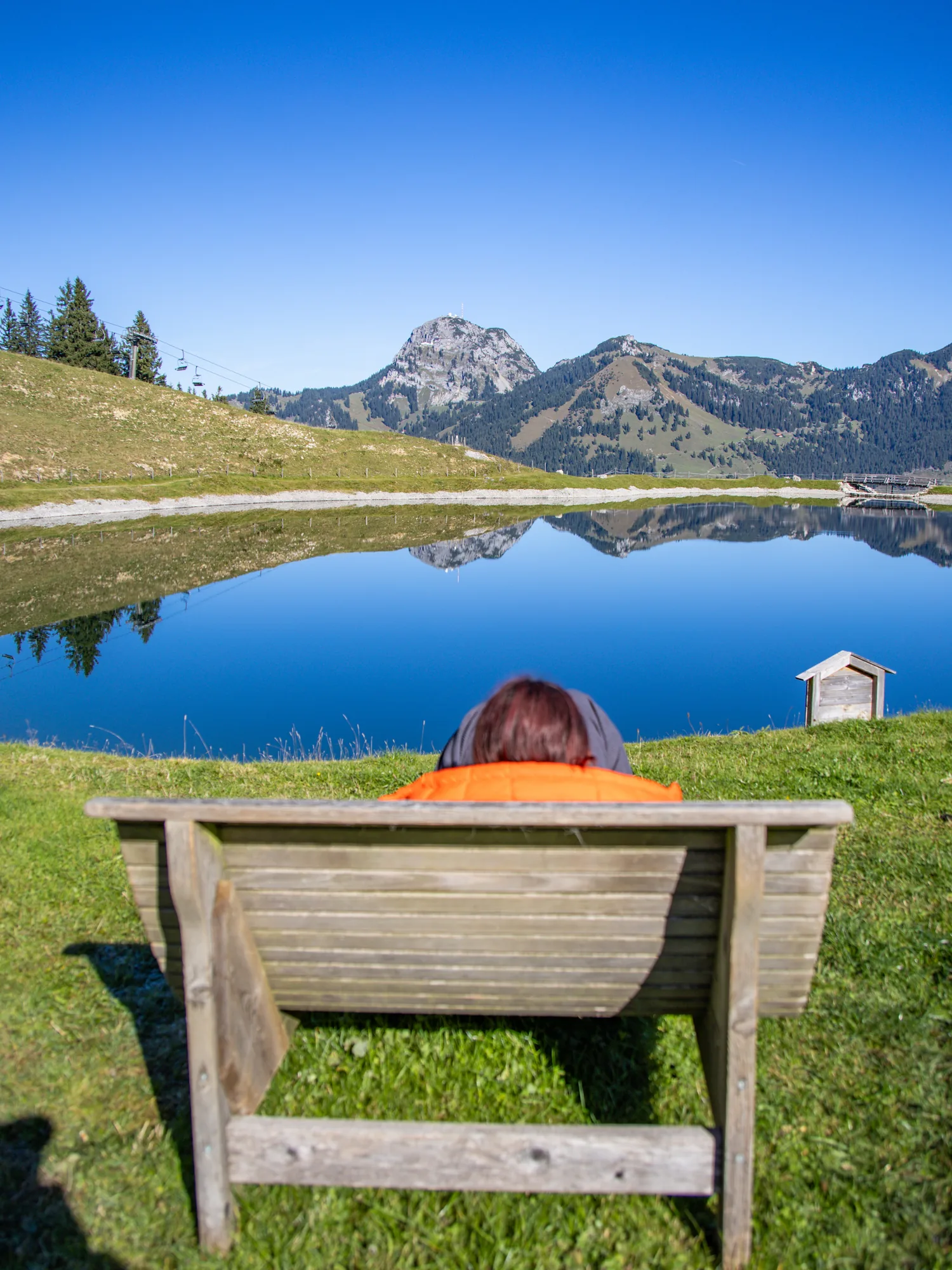 Person auf Bank an einem Bergsee mit Spiegelung der Gipfel