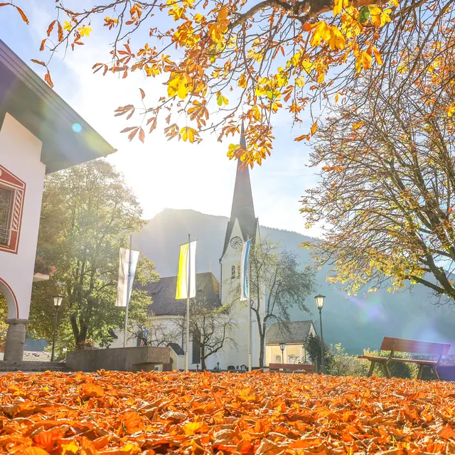 Kirchturm hinter herbstlich gefärbtem Baum mit Laub am Boden